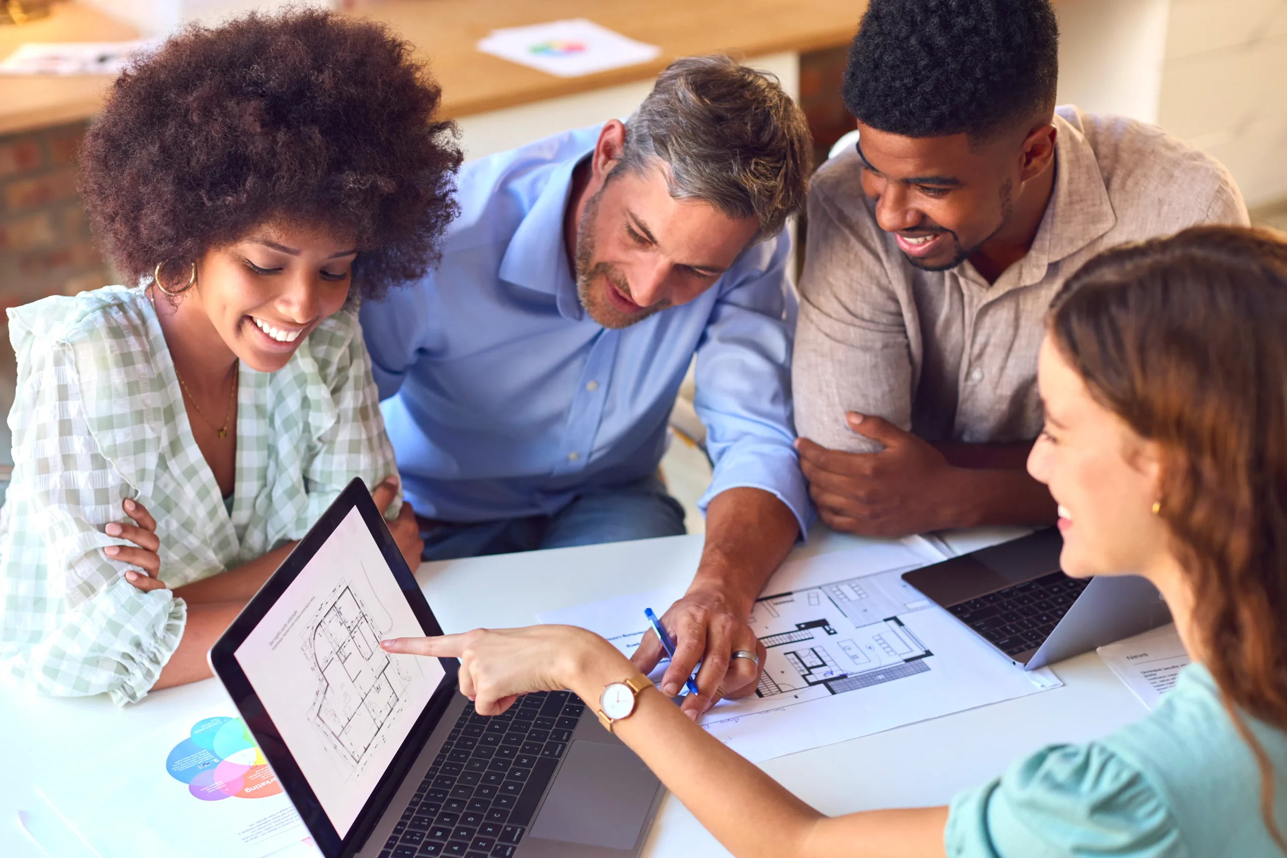 Quatro profissionais estão reunidos em uma mesa de trabalho. Uma mulher de cabelos cacheados observa a tela de um notebook, onde outra mulher, à frente, aponta para um esquema de planta apresentado. Ao lado, dois homens acompanham atentamente a explicação. Sobre a mesa há papéis, laptops e documentos, transmitindo a ideia de colaboração e planejamento estratégico. A cena representa a atuação da Kodevo em consultoria web, destacando o trabalho em equipe e a análise de soluções personalizadas para os clientes.