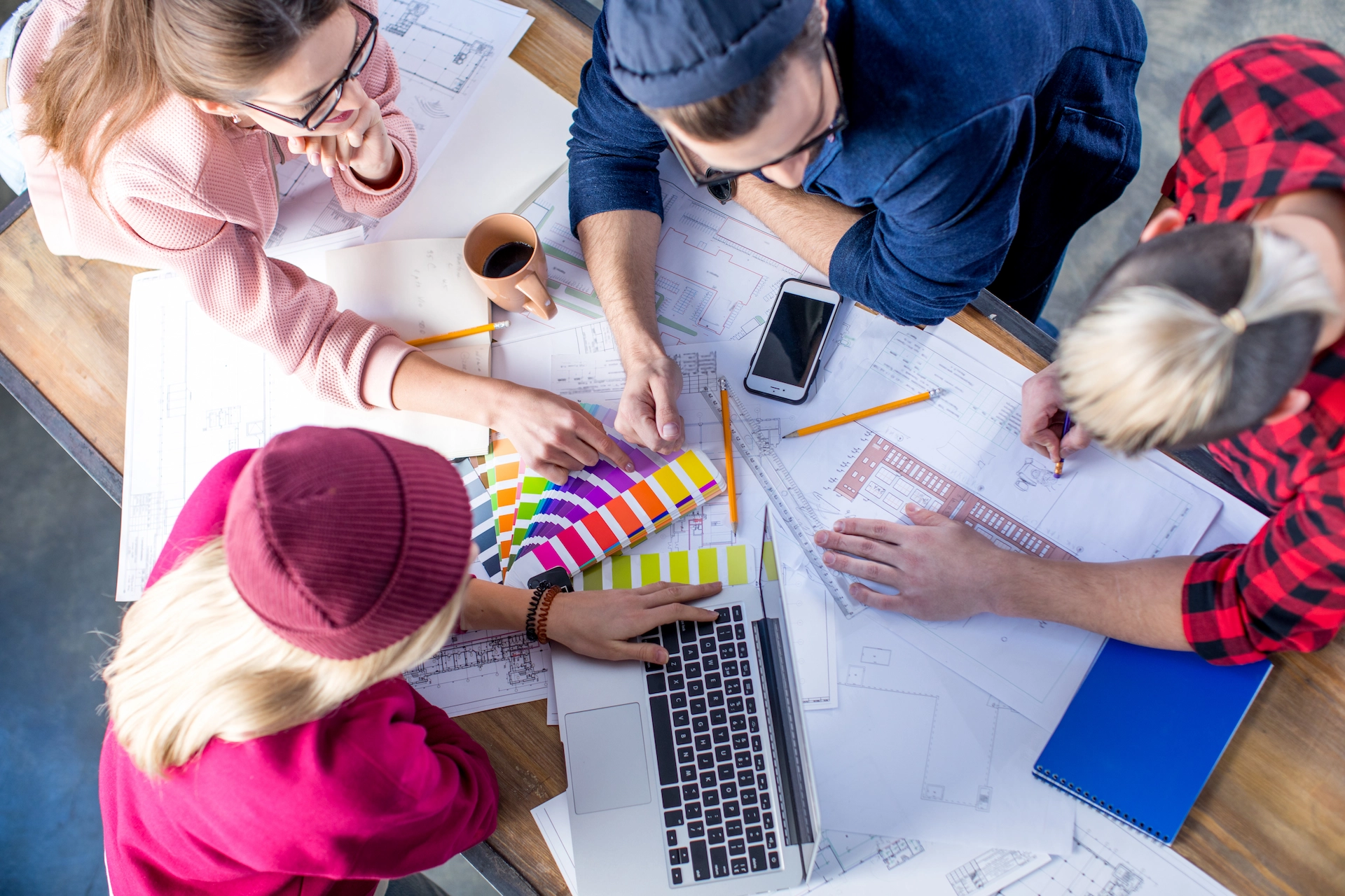 Um grupo de profissionais trabalha em conjunto sobre uma mesa repleta de plantas, anotações e amostras de cores. No centro, um notebook está aberto e todos discutem ideias e possibilidades criativas. A cena transmite colaboração e inovação, sendo uma representação clara de processos ligados ao web design, onde conceitos visuais e funcionais são desenvolvidos para criar experiências digitais atrativas e eficientes.